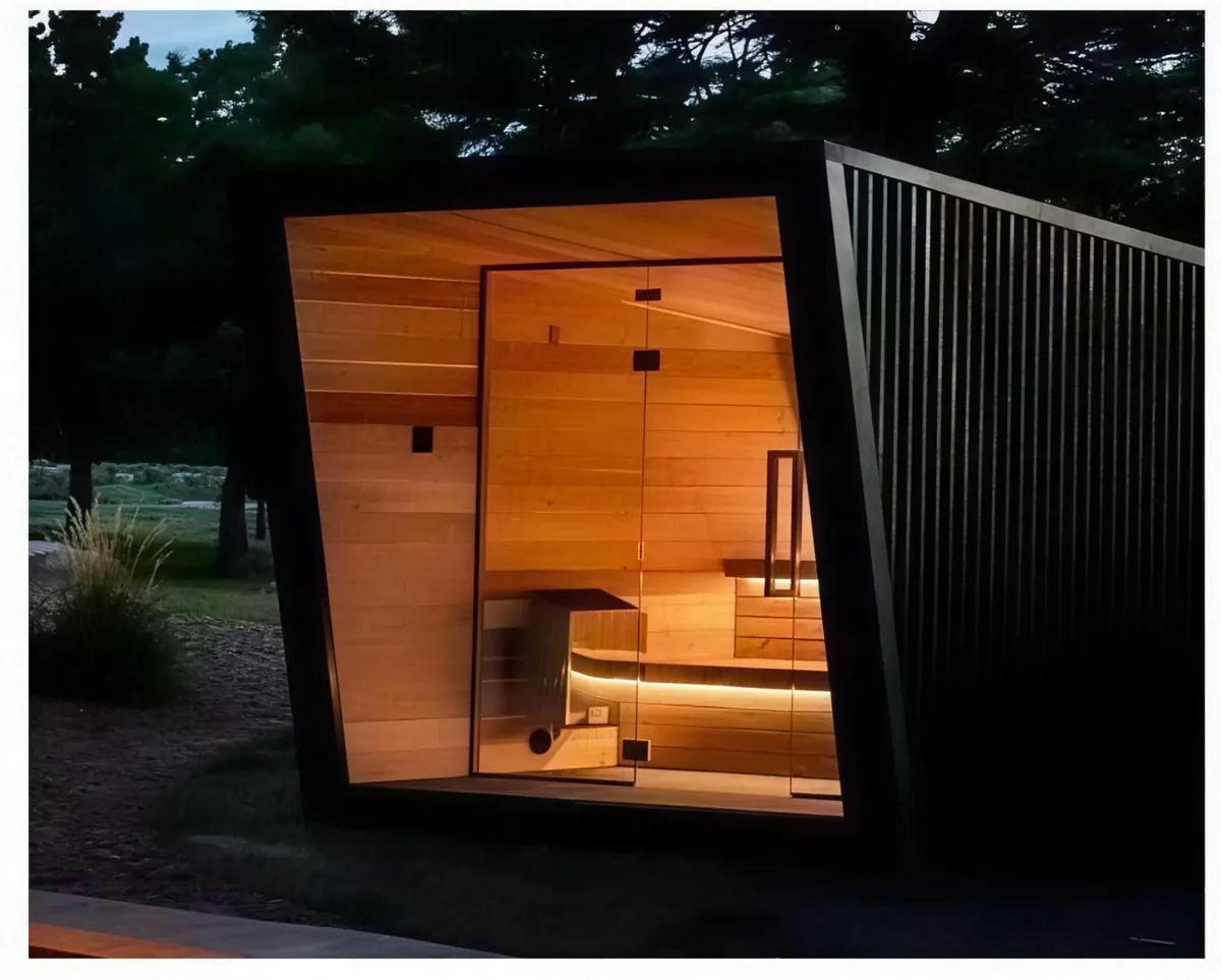 Kabin outdoor sauna bentuk khas dengan fasad hitam condong dan dalaman hemlock yang bercahaya pada waktu senja
